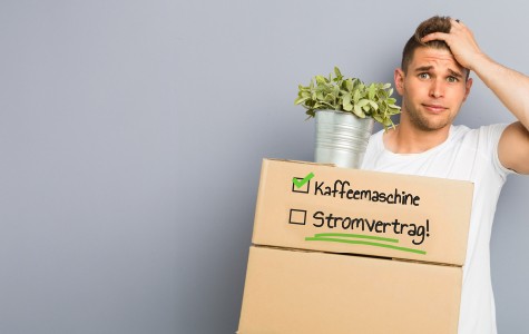 Ein junger Mann steht vor einer grauen Wand und hält zwei Umzugskartons sowie eine Topfpflanze in den Händen. Auf den Kartons stehen handgeschriebene Notizen mit Häkchen neben "Kaffeemaschine". Auf dem unteren Karton ist "Stromvertrag!" mit einem leeren Kästchen geschrieben. Der Mann schaut besorgt und kratzt sich am Kopf – möglicherweise, weil er vergessen hat, den Stromvertrag abzuschließen.